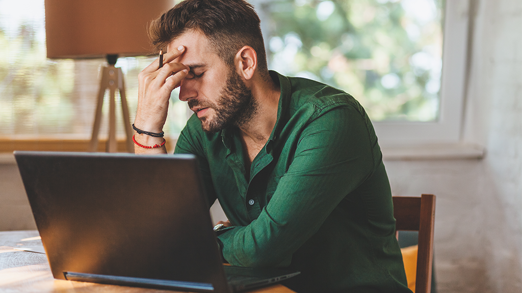 A young man sitting on a chair, his laptop on the desk, is thinking with his eyes closed, tired and stressed