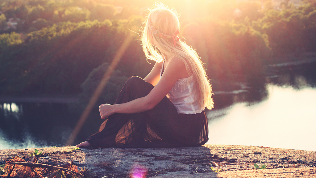 A young blonde woman, turning her back, looking at the sea sitting on the top of a hill 